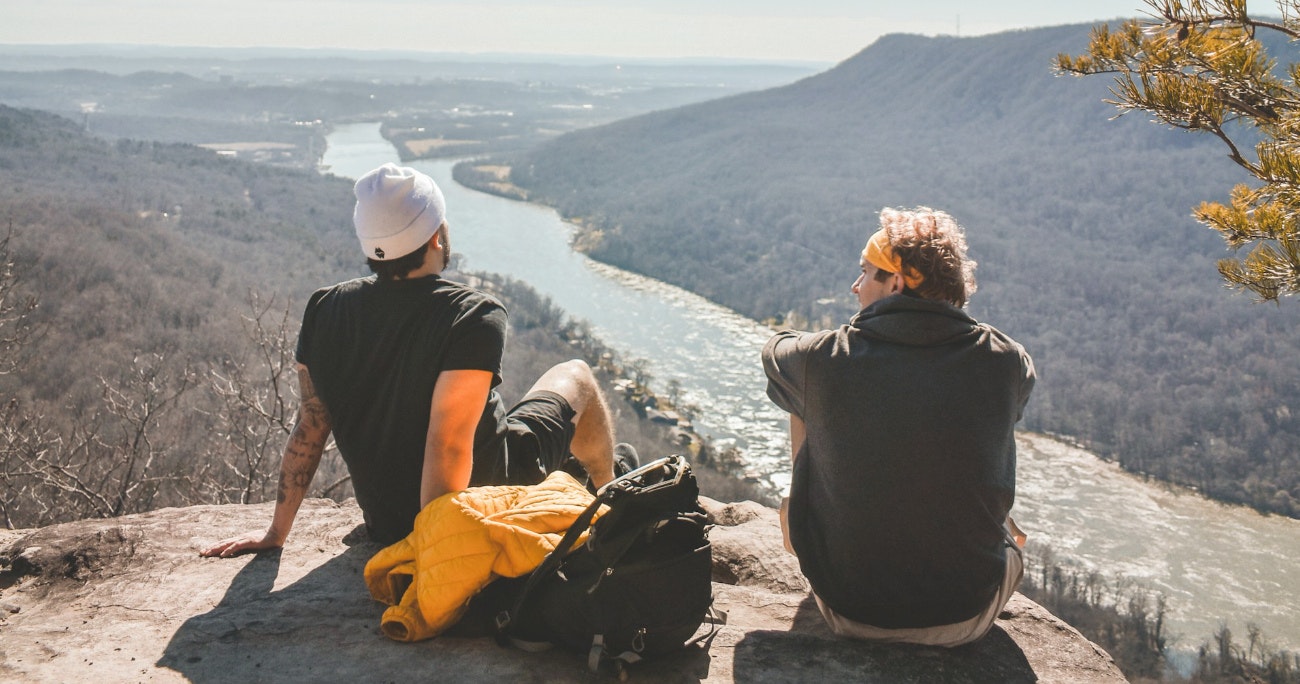 two guy sitting and talking overlooking a river in a valley below